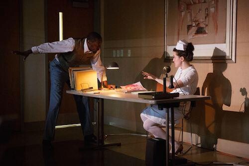 On stage, a woman is sitting at a desk and a man is leaning over it and pointing away.
