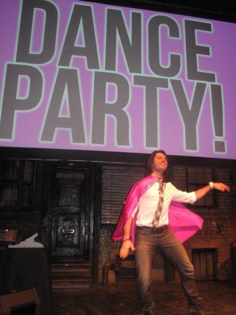 A person on stage in a pink cape with a screen behind them that says Dance Party.