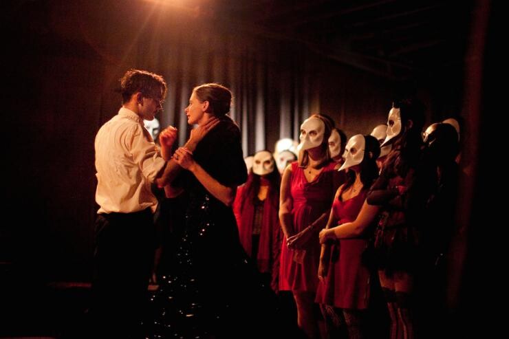 A shot of a stage production with two people clutching each other and a group of masked onlookers.