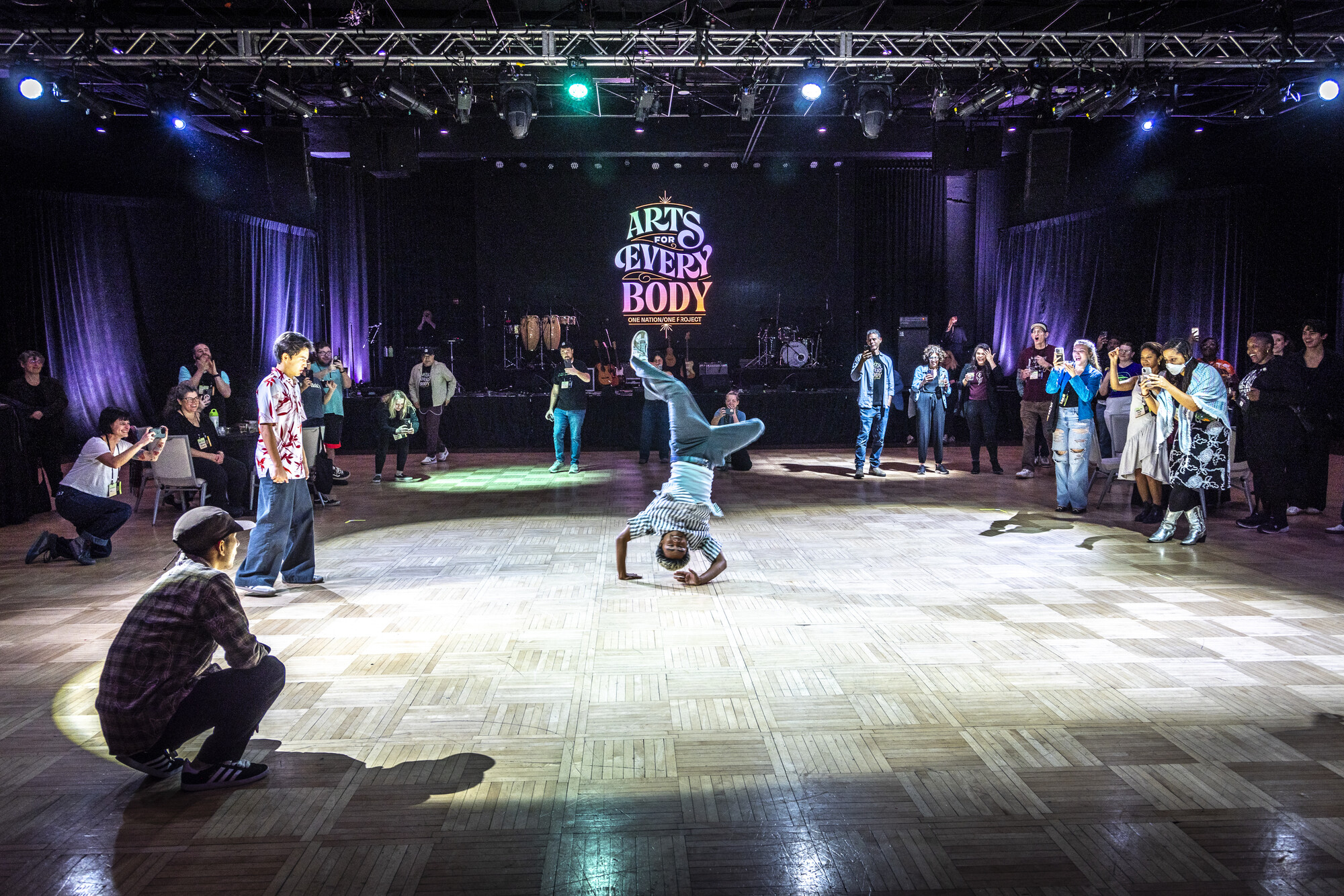 People standing round the edges of a dance floor watching a person in the center break dance.