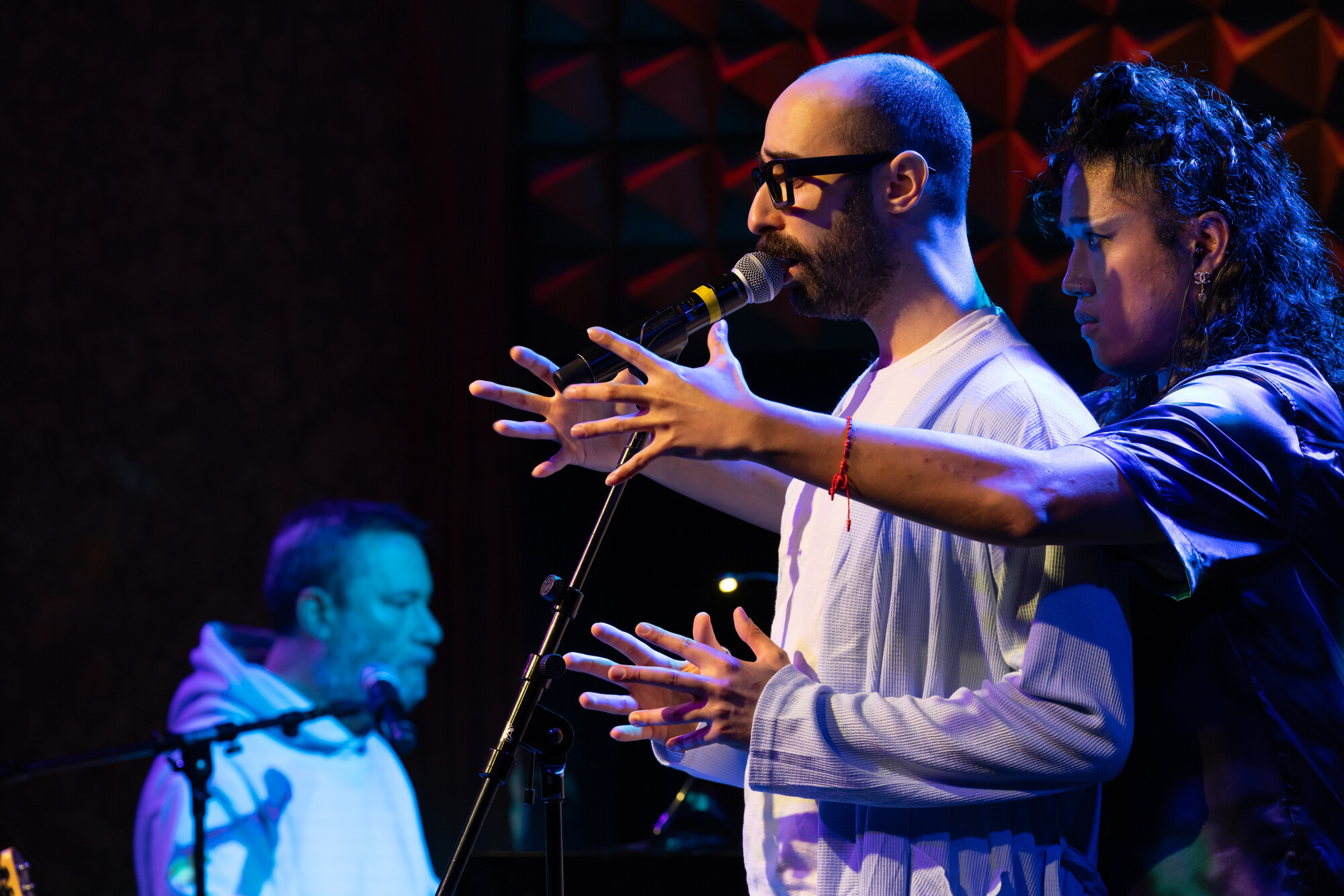 A person singing on stage, with another person behind reaching forward.