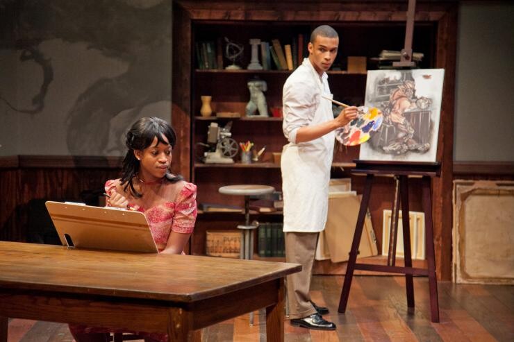 A man painting on a cavas on stage and a woman sitting at a table.