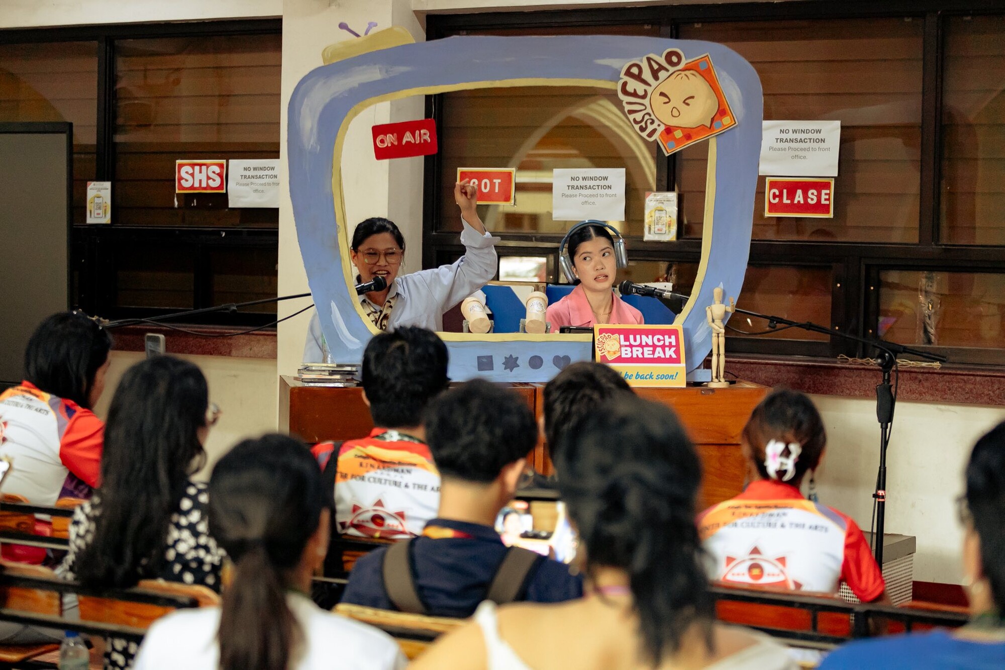 An audience watching two people in a cardboard news tv set.