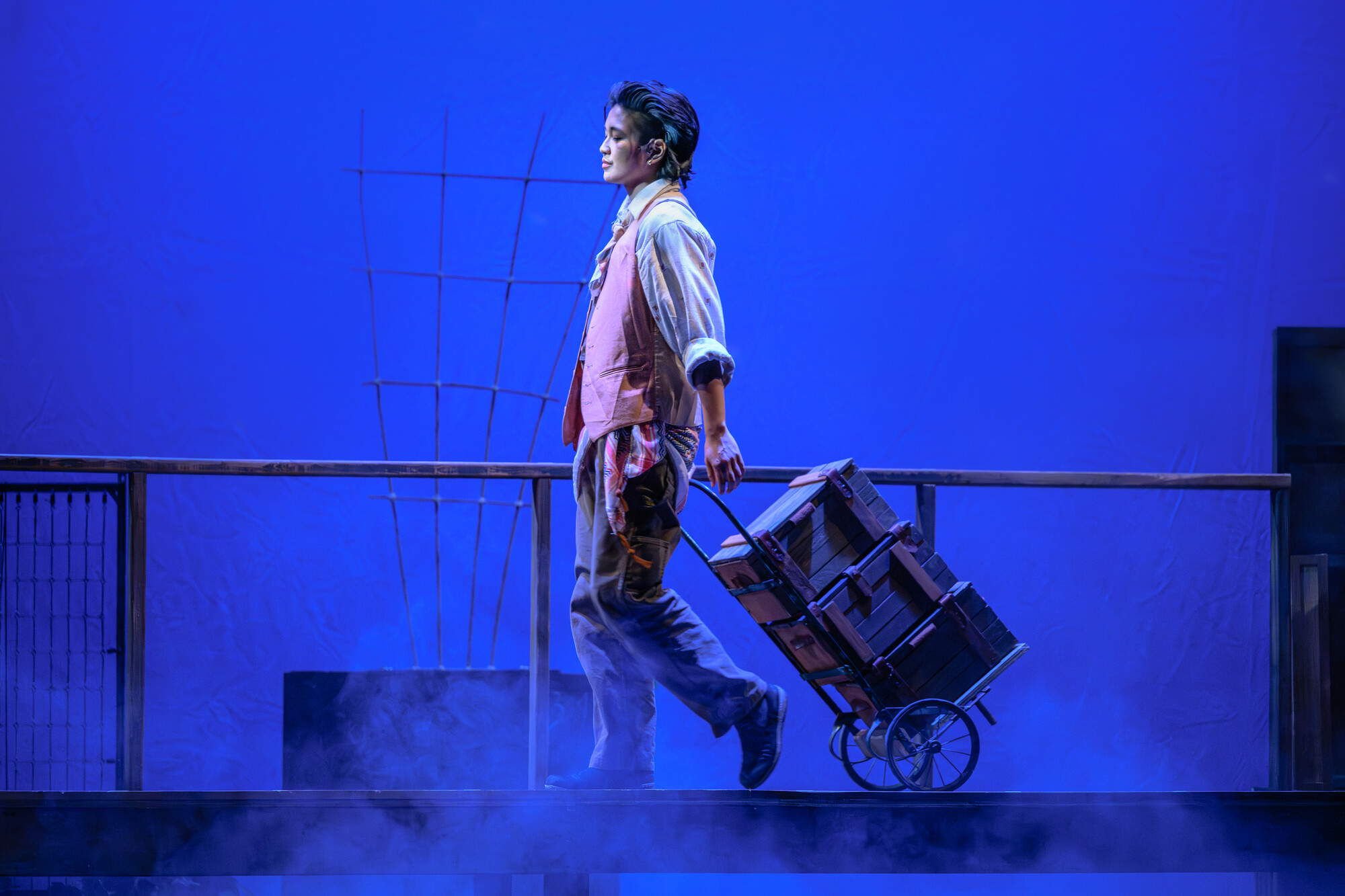 An actor on stage pulling a cart with three boxes on it.