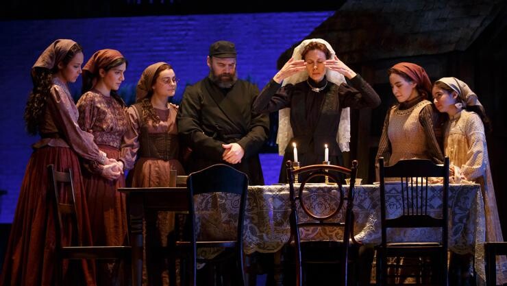 A group of actors standing near a table set for dinner on stage.