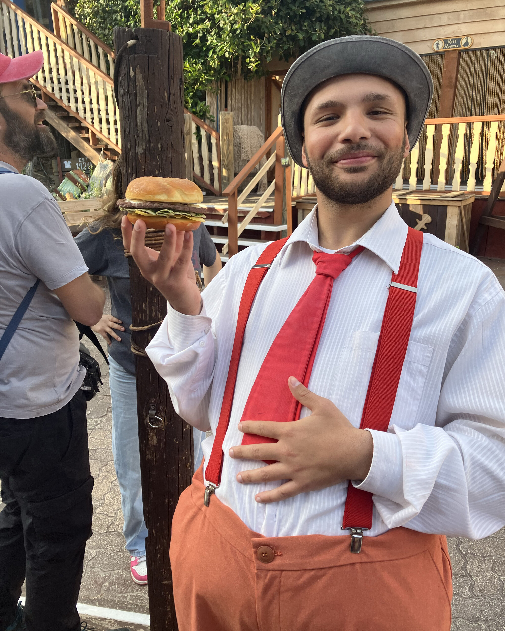 A man in a a white shirt, orange pants, red tie, and red suspenders holding a burger.