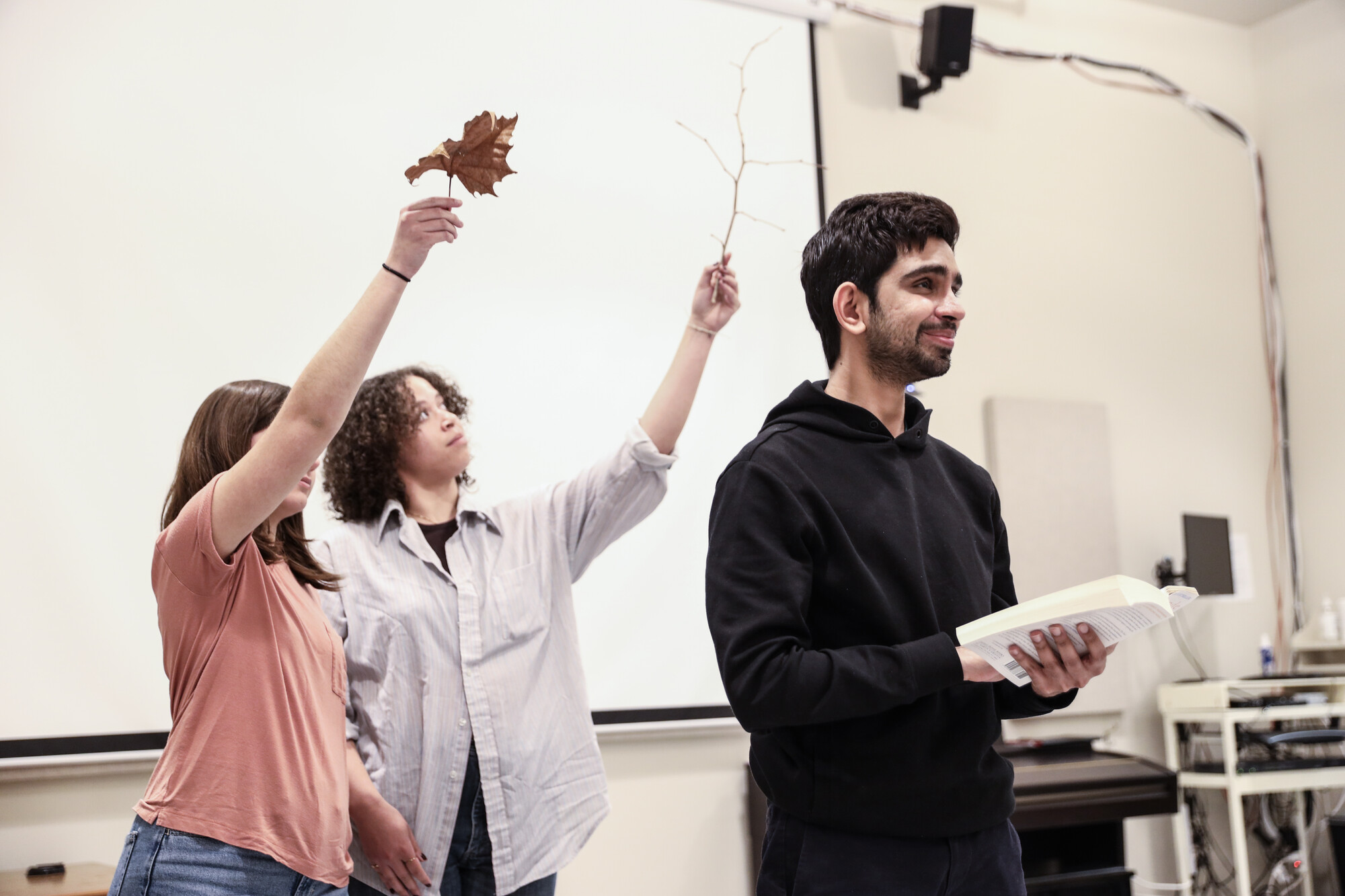 Three people in a rehearsal room, two holding leaves over the head of another.