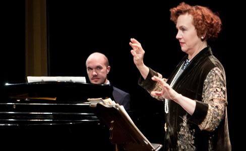 A man at the piano and a redhead woman conducting.