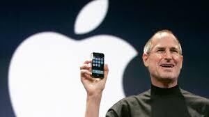 Steve Jobs holding up an old iphone standing in front of a giant white Apple logo.