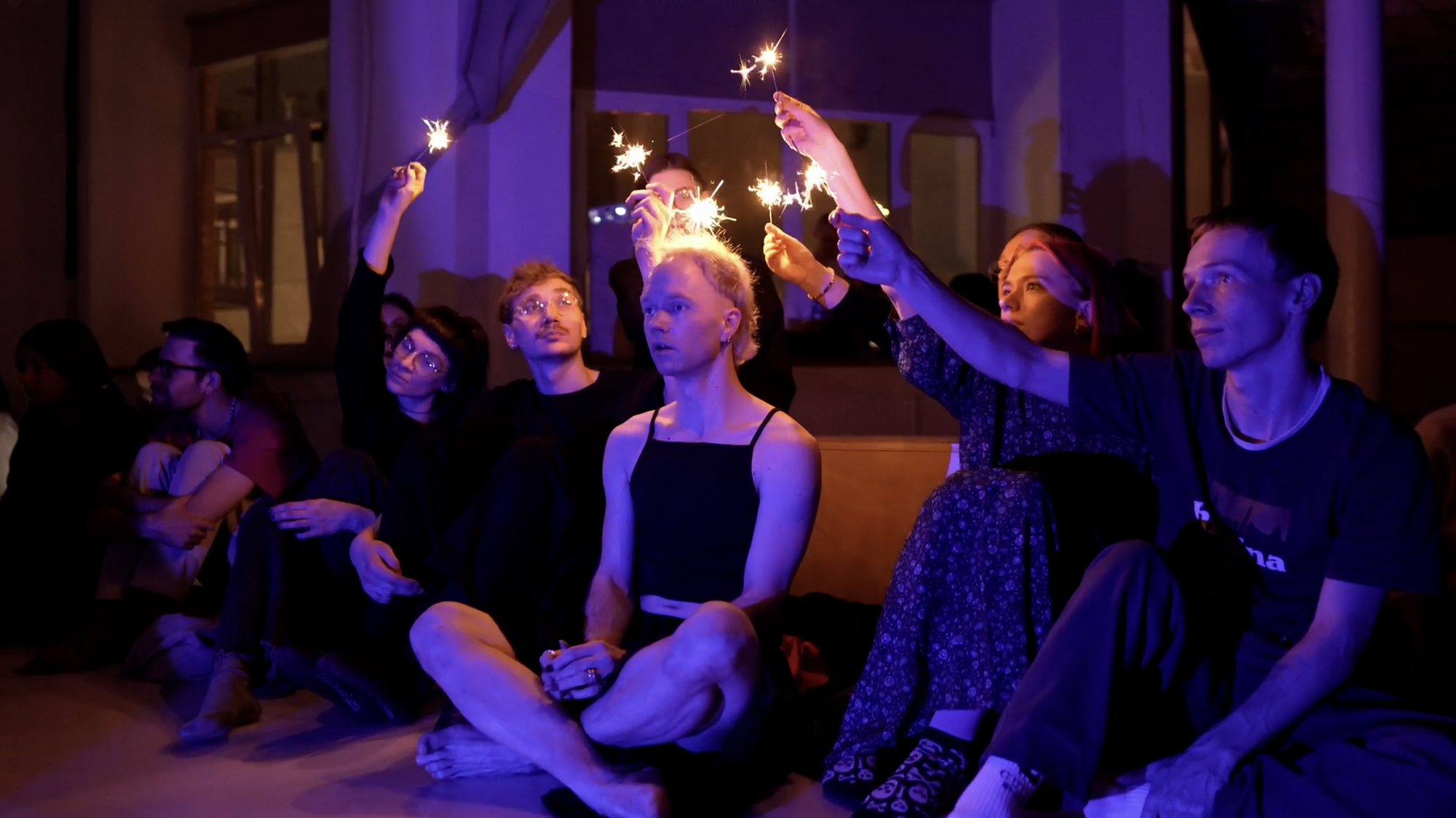 A group of people sitting on the floor holding sparklers over the head of someone else.