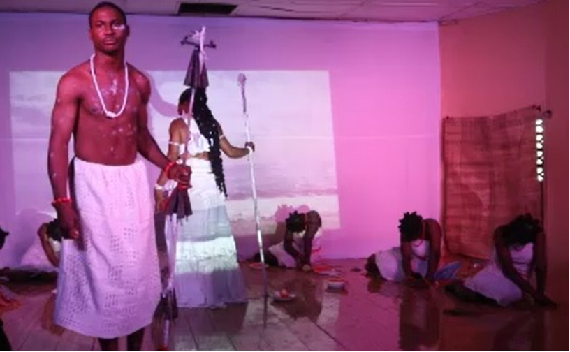 A man in a skirt holding a spear on stage while three women are on the floor kneeling.