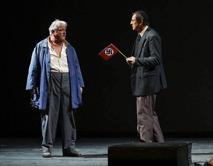 Two men on stage, one in a slouchy blue jacket and the other holding a Nazi flag.