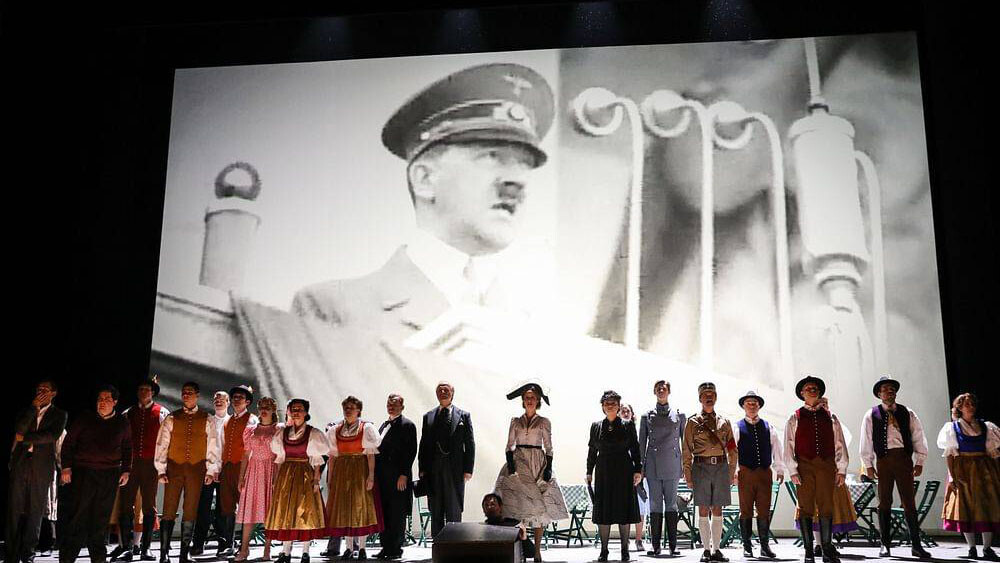 A row of actors standing on stage with a photo of Hitler behind them on the screen.