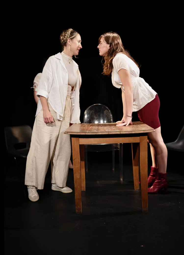 Two women on stage standing up and looking at each other over a table.
