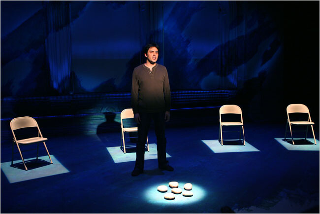 A person standing alone on stage in front of four metal folding chairs.