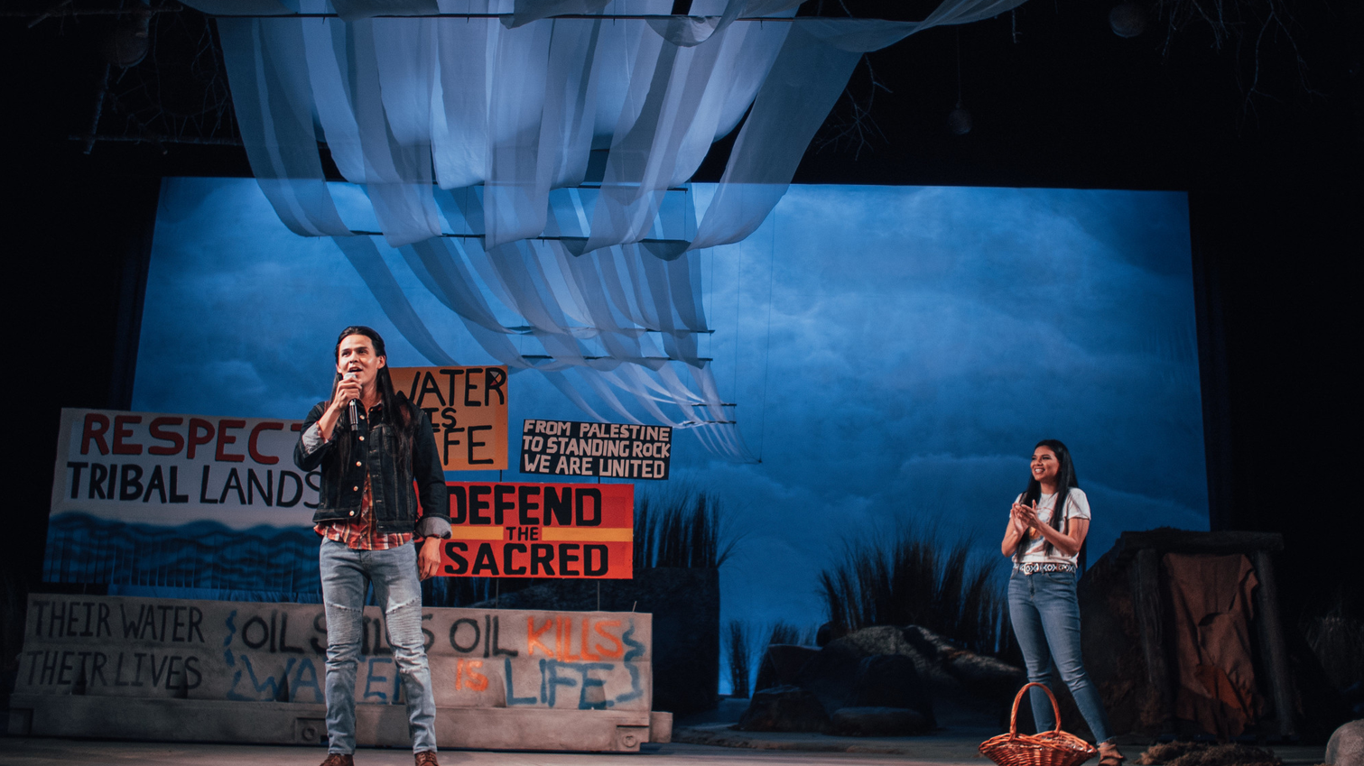 Two actors on stage with lots of signs in the background.