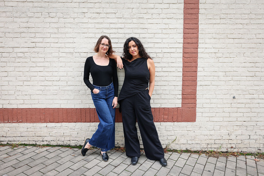 Two women dressed in black taking a picture in front of a white wall with a red stripe.