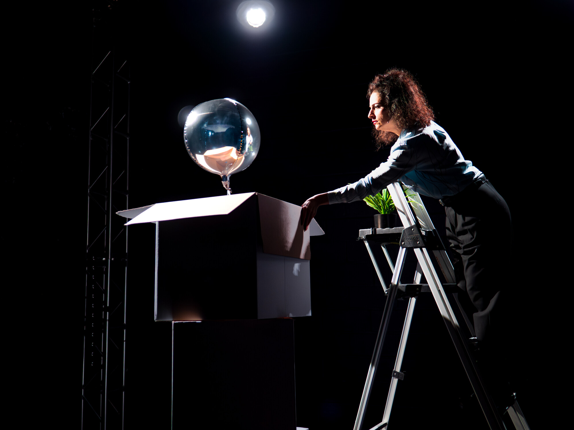 A woman on stage on holding on to a cardboard box with a silver ball on the top.