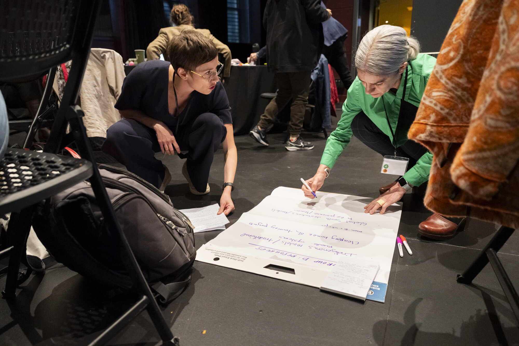 Two women writing on a piece of paper on the ground.