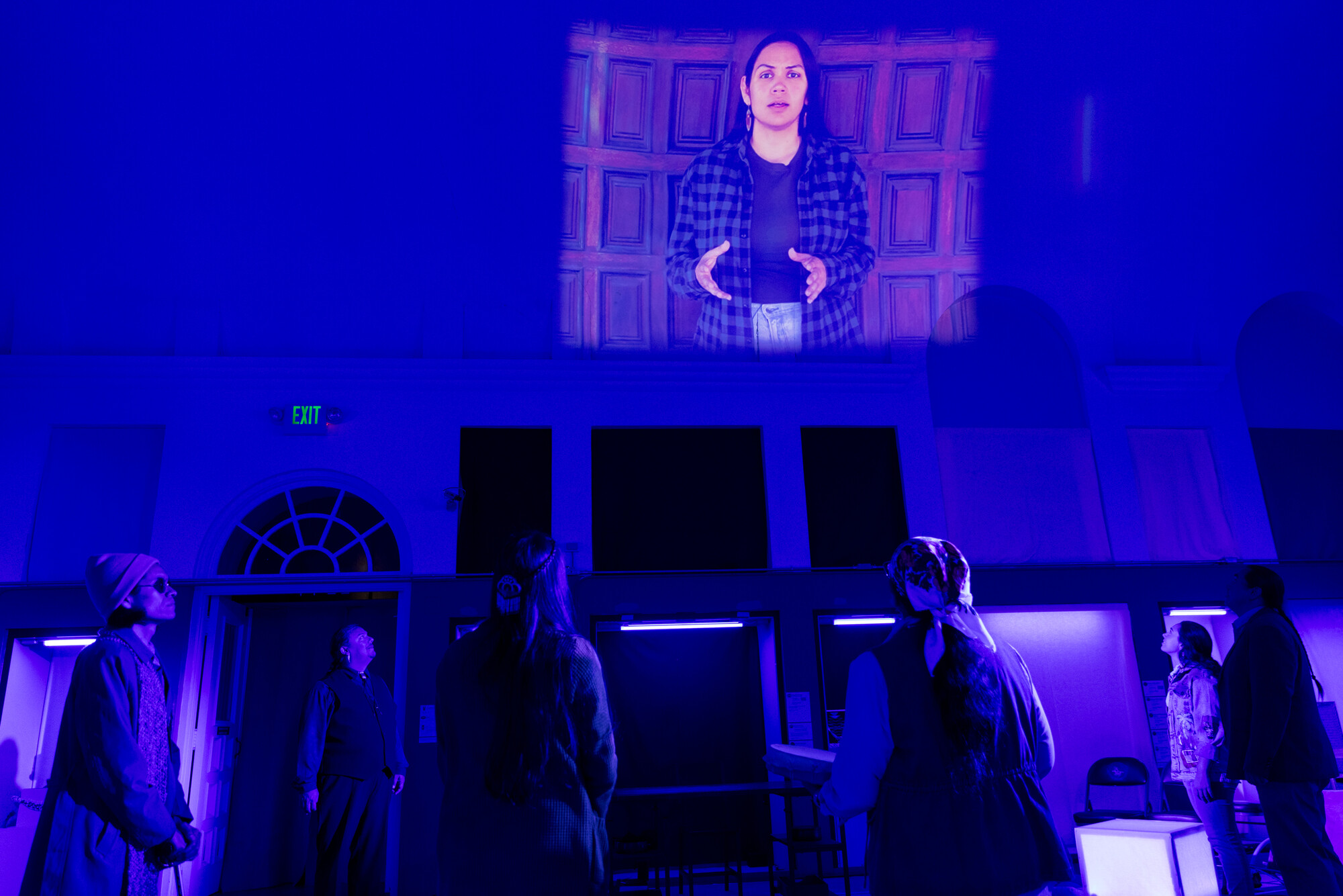 A group of people onstage look up to a projector screen of a person speaking.