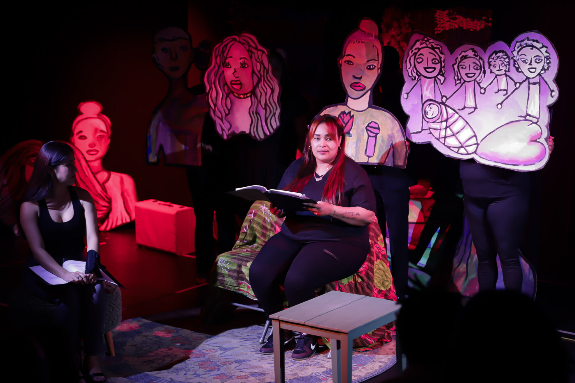 Two people sitting on stage reading with puppets in the background.