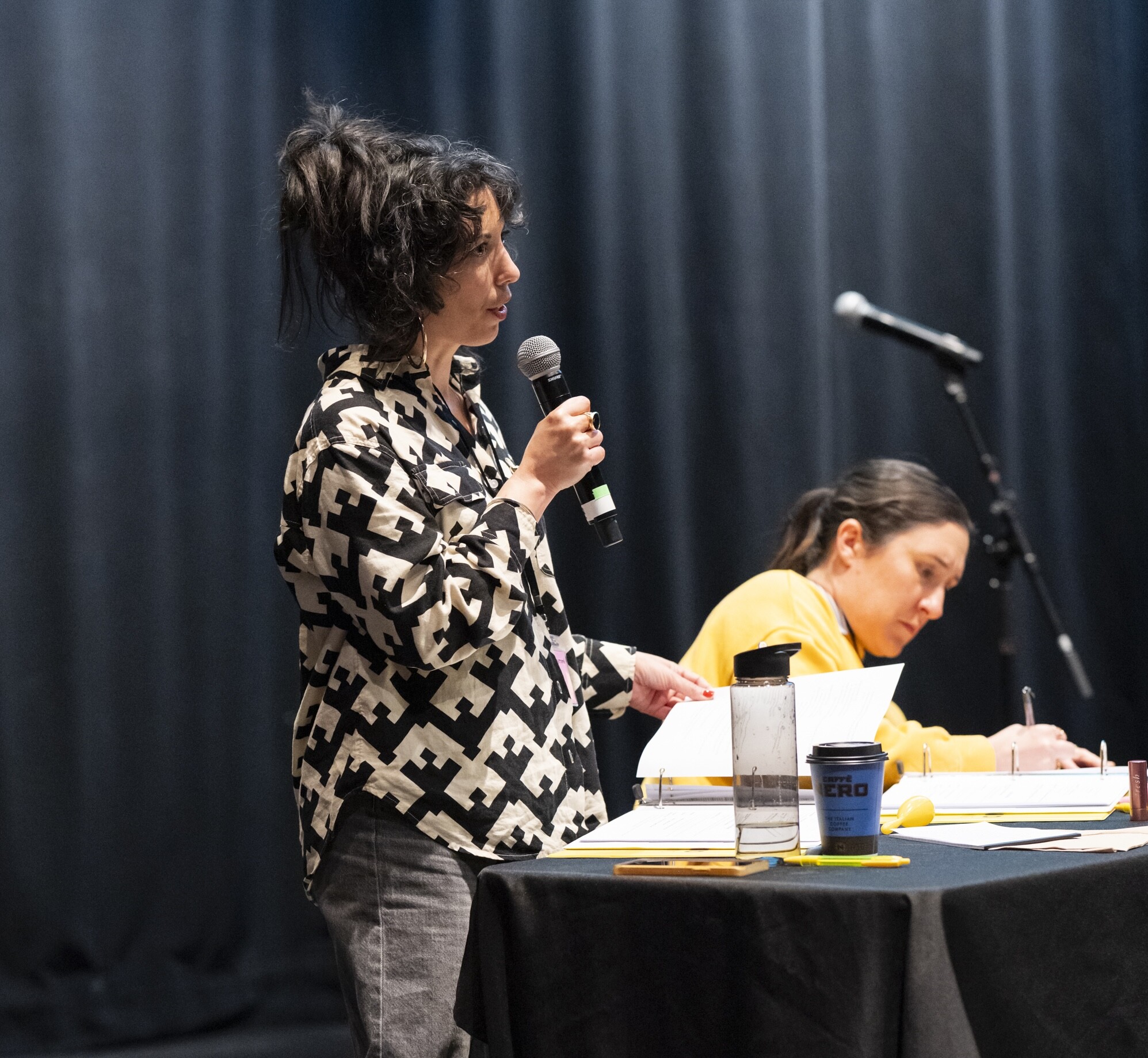 A woman with dark hair speaks to a room on a microphone.