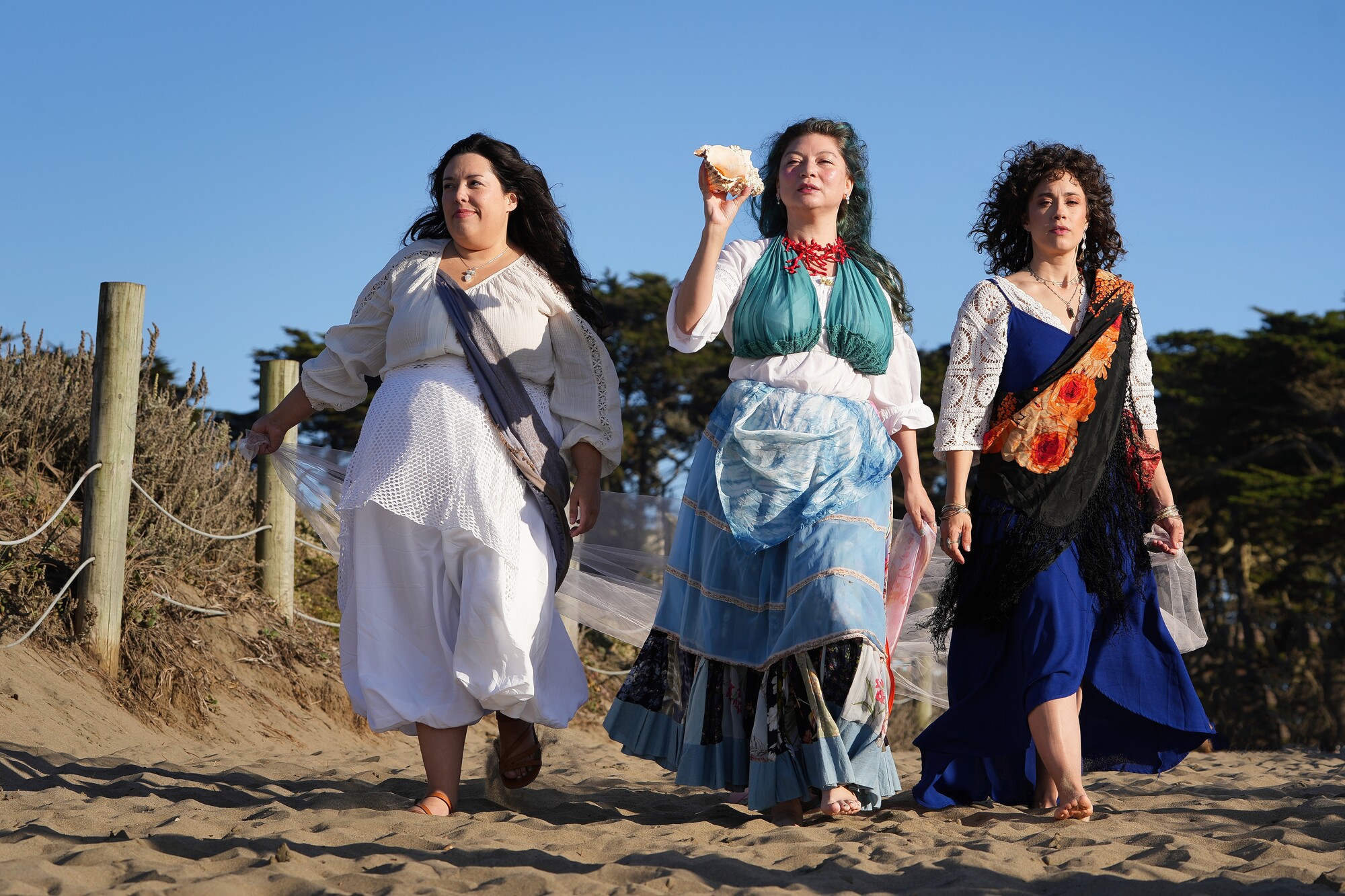 Three women in long dresses walk on a beach.