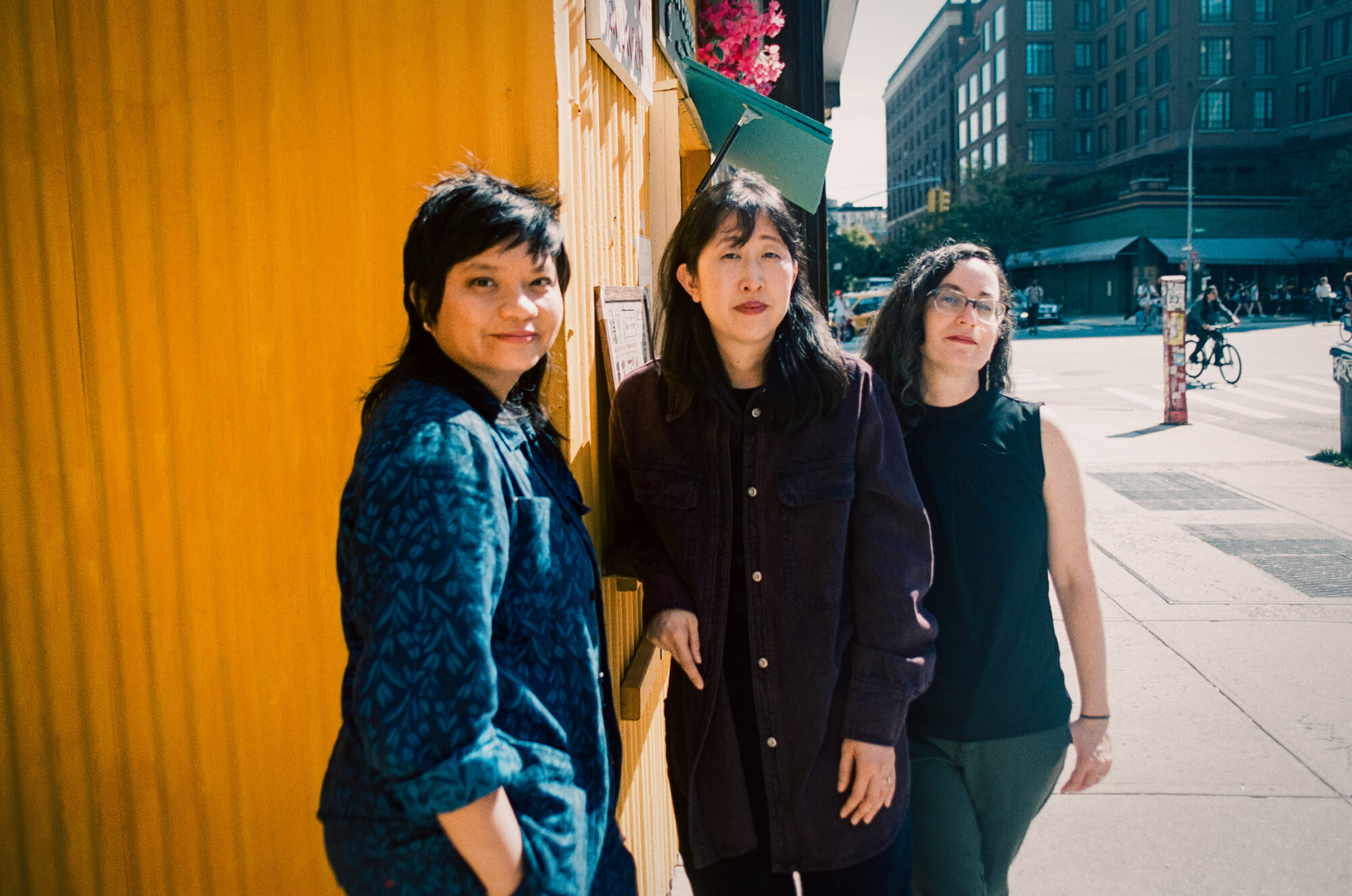Three people lean against a wall on a city street.
