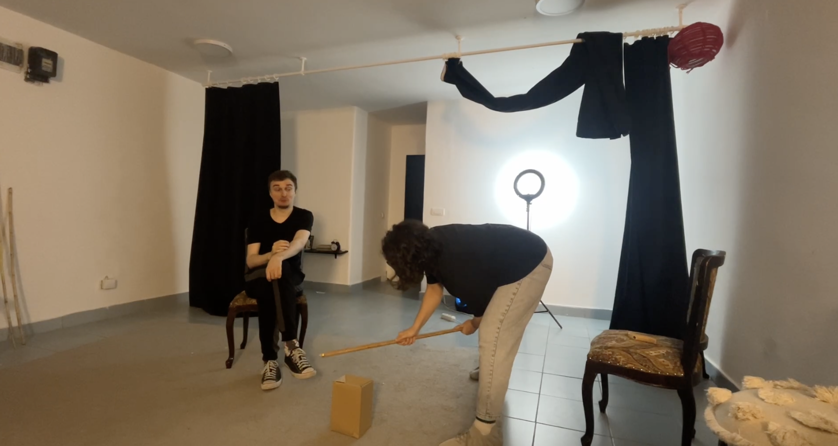 Two actors move props in a small white room.