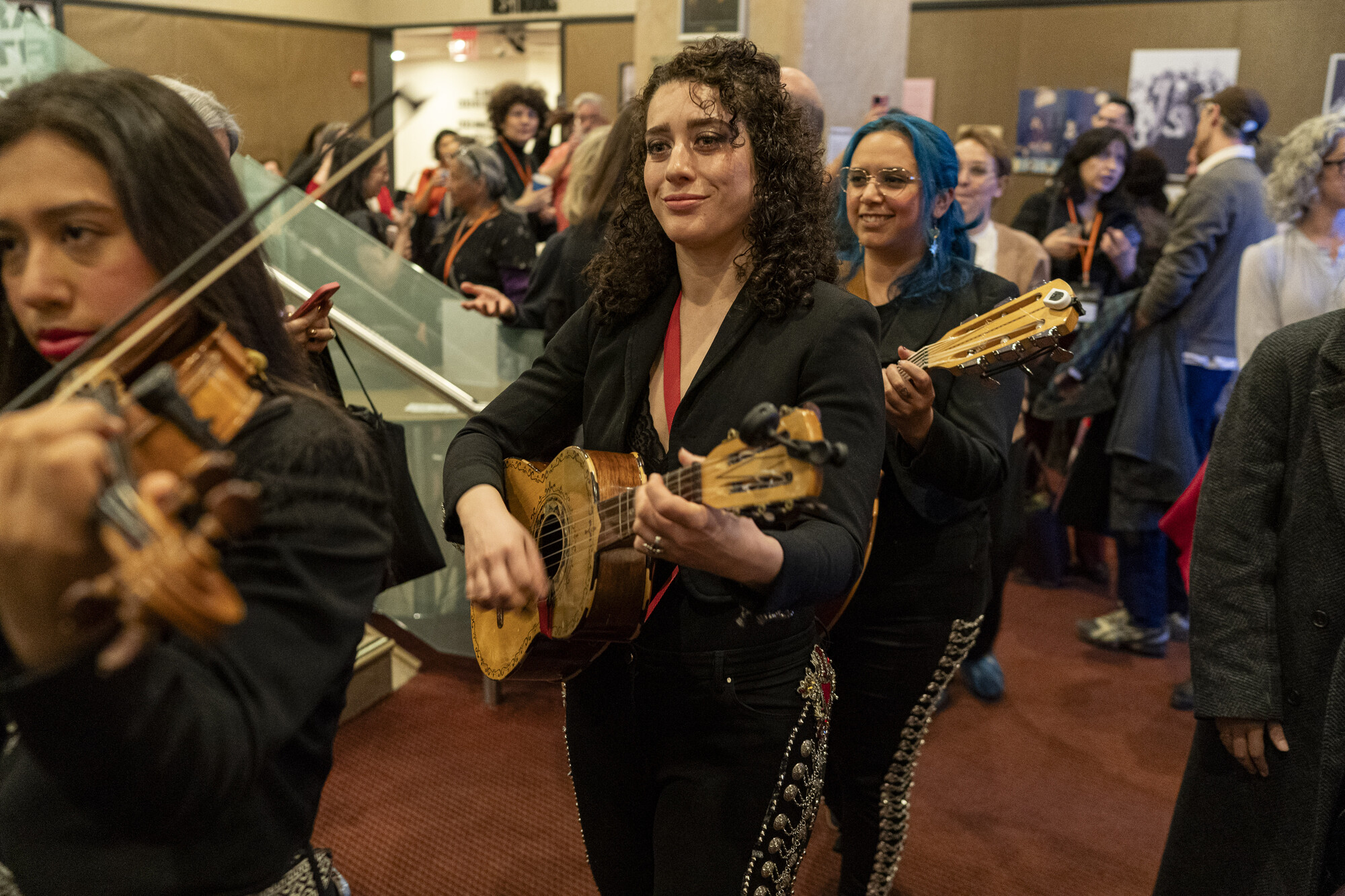 A row of women playing instruments walk through a crowd.