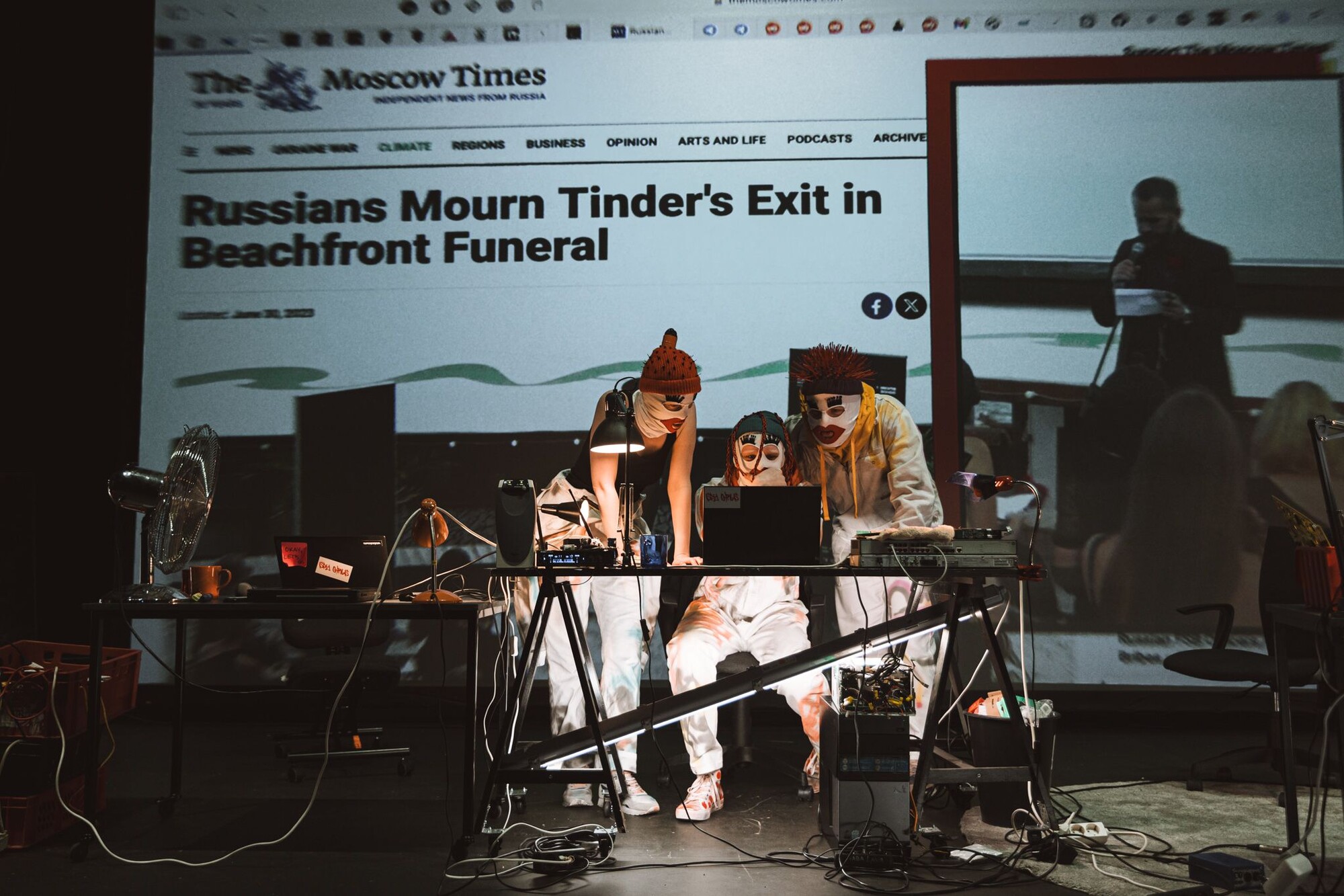 Three people in masks lean over a laptop onstage.