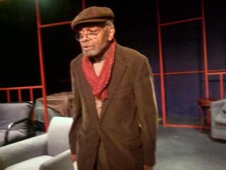 A older black man in a brown coat, red scarf, and a brown newsboy cap.