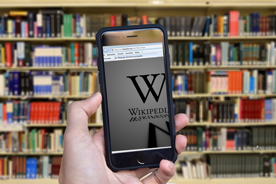 A person holding up a phone displaying a Wikipedia page in front of a library shelf.