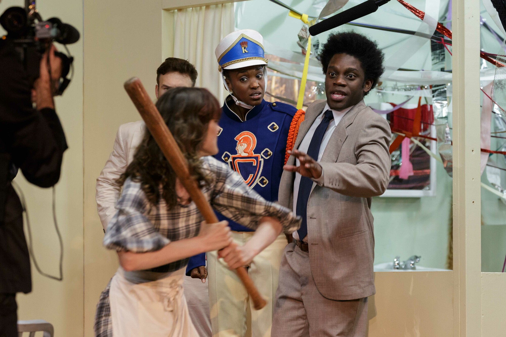 Several actors onstage. A woman swings a baseball bat at two others.