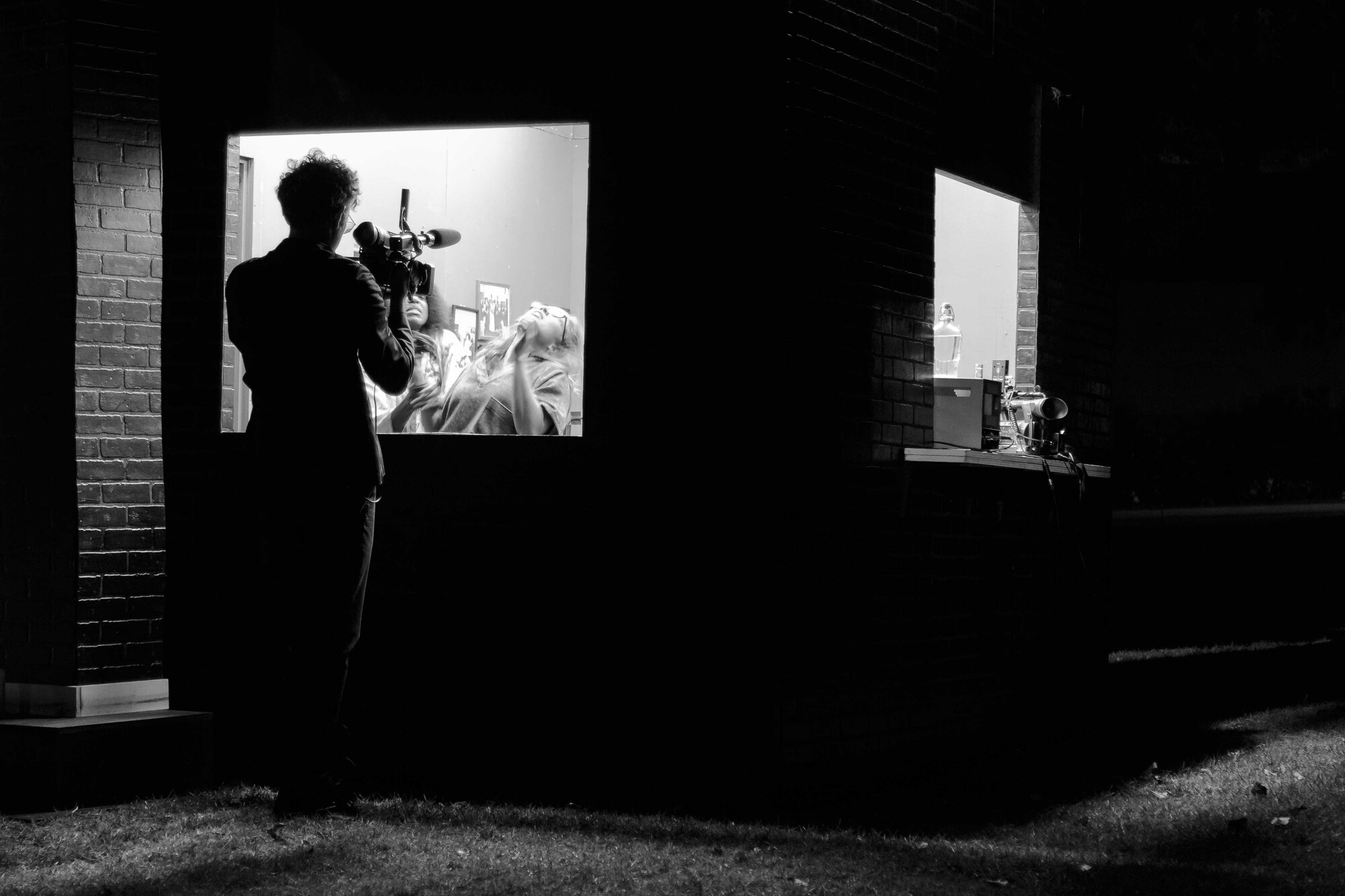 A man in silhouette films two actors performing behind a screen.