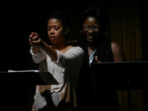 Two Black performers, one with their arms extended, stand in front of music stands.