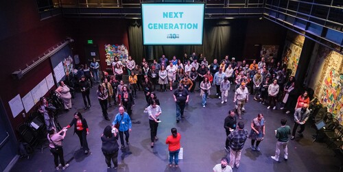 A group of people walk across a black box theatre space under a sign saying Next Generation