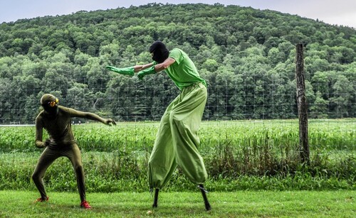 two figures in green, one on stilts, fight in a field